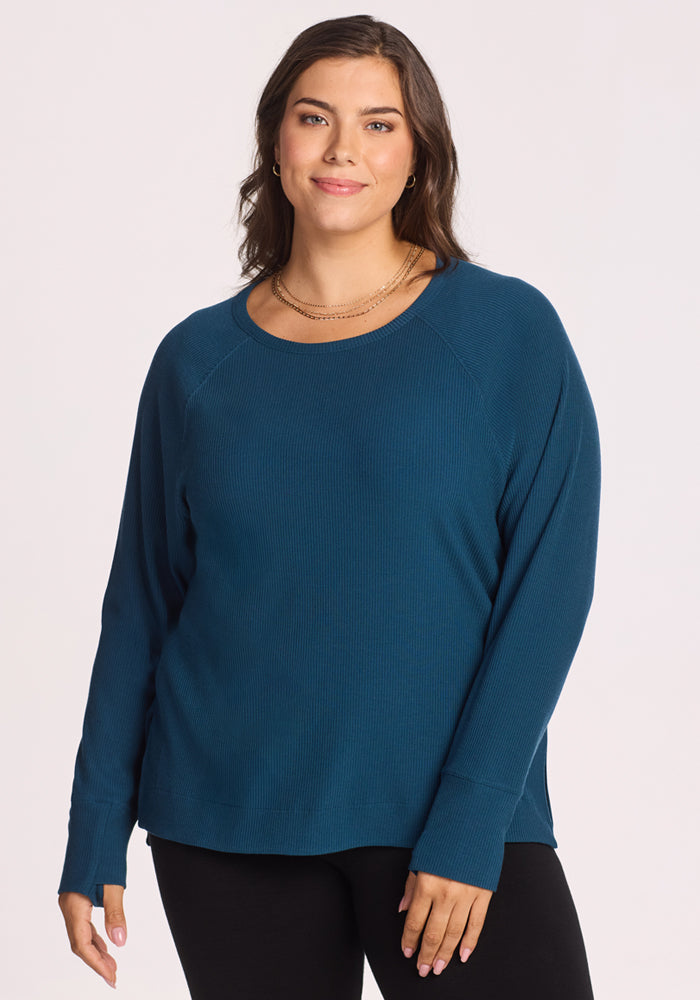 A woman with long brown hair wears the Woolx Ainsley Sweater in Majolica Blue and black pants, standing against a plain light background and smiling gently at the camera. 
