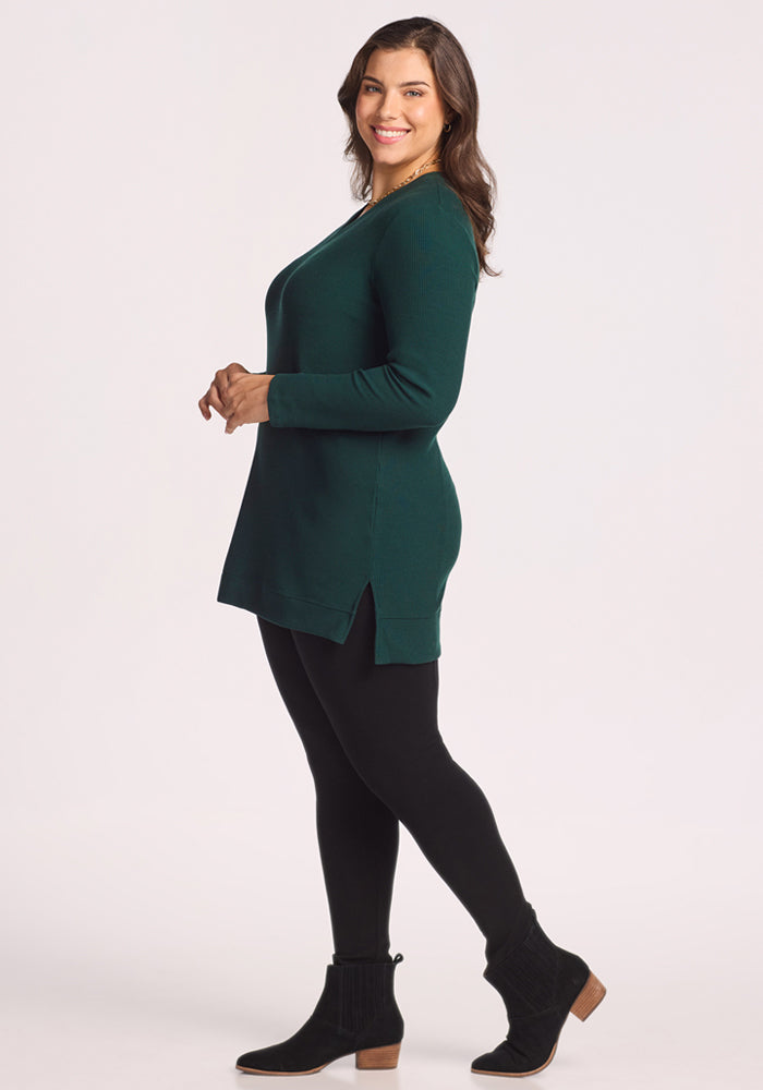 A woman stands sideways, smiling, wearing the Woolx Evie Ribbed Tunic in Deep Timber with black leggings and low-heeled black ankle boots, set against a plain light background.