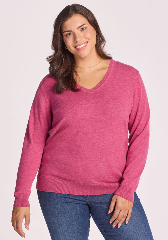 A woman with long brown hair, wearing the Woolx Francesca Sweater in Wild Salmon and blue jeans, smiles while standing against a plain light background. 