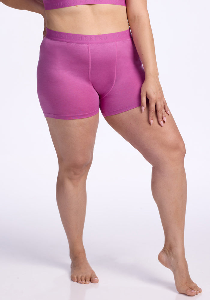 A person stands barefoot on one leg against a plain white background, wearing Woolx’s Elliott Boyshort - Wild Blossom and a matching pink sports bra. Their left hand rests gently on their thigh. 