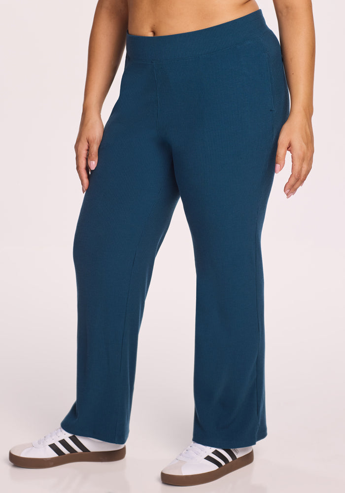 A person wearing Woolx Luca Ribbed Pants in Majolica Blue and white sneakers with black stripes stands against a plain light background, showing only the lower half of their body. 