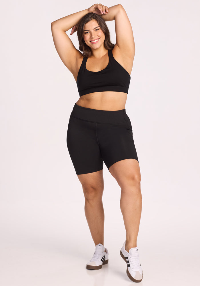 A woman in a black sports bra, Woolx Merino Flex® Dani Bike Shorts - Black, and white sneakers stands smiling with one arm raised behind her head against a plain white background.