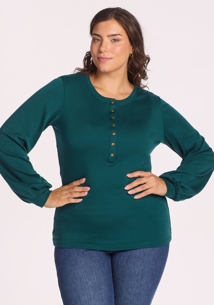 A woman with long brown hair wears the Woolx Charlotte Puff Sleeve Top in Riverbank Green and blue jeans, standing with one hand on her hip against a plain white background. 