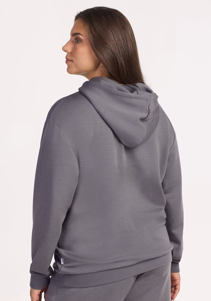 A person with long brown hair wearing the Woolx Avery Hoodie in Arctic Dusk and matching pants stands facing away from the camera, looking to the side against a plain, light-colored background, ideal for year-round comfort.