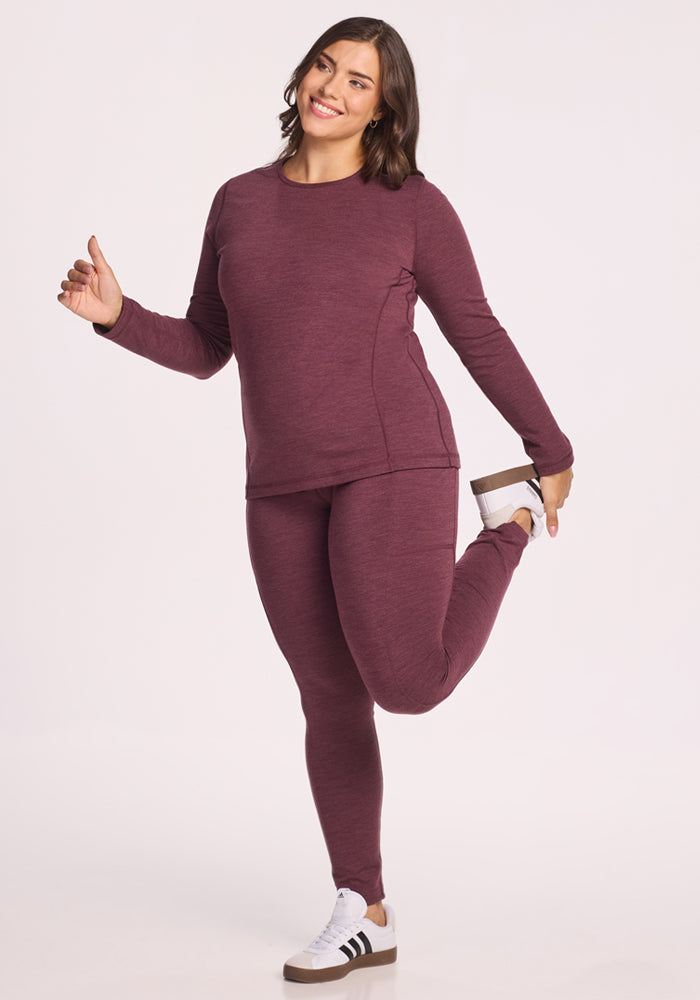 A woman wears the Woolx Elsa Long Sleeve in Grape Harvest Melange, smiling and stretching on one leg against a plain light background.