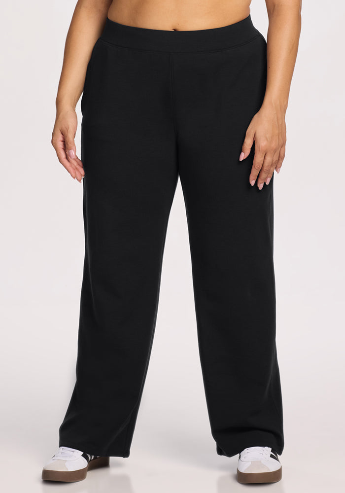 A person wearing Woolx Brynn Pants in Black and white sneakers stands with hands relaxed by their sides against a plain light background. The upper body is partially visible. 