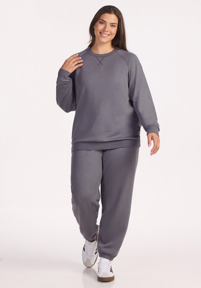 A woman with long brown hair smiles while walking, wearing the Woolx Bailey Crewneck Sweatshirt in Arctic Dusk and matching gray sweatpants with white sneakers, against a plain white background.