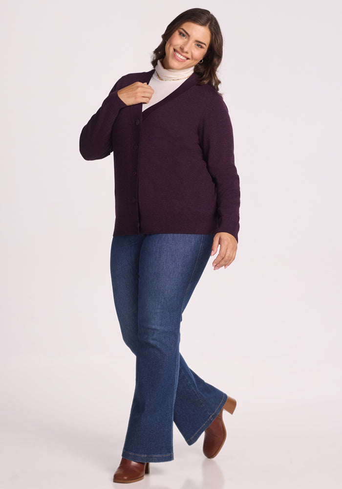 A woman with shoulder-length brown hair smiles while wearing the Woolx Gia Sweater in Deep Plum, paired with a white turtleneck, blue flared jeans, and brown boots, standing against a white background.