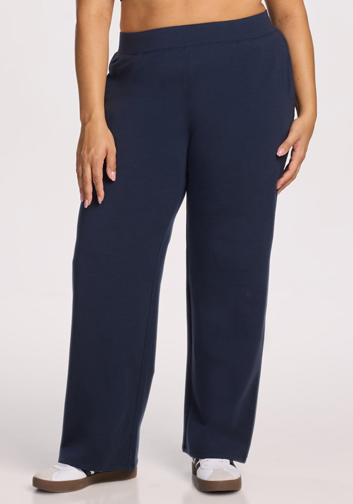 A person wears Woolx Brynn Pants in Deep Navy, styled wide-leg in merino wool, paired with white sneakers against a light background. Only the lower torso and legs are visible.