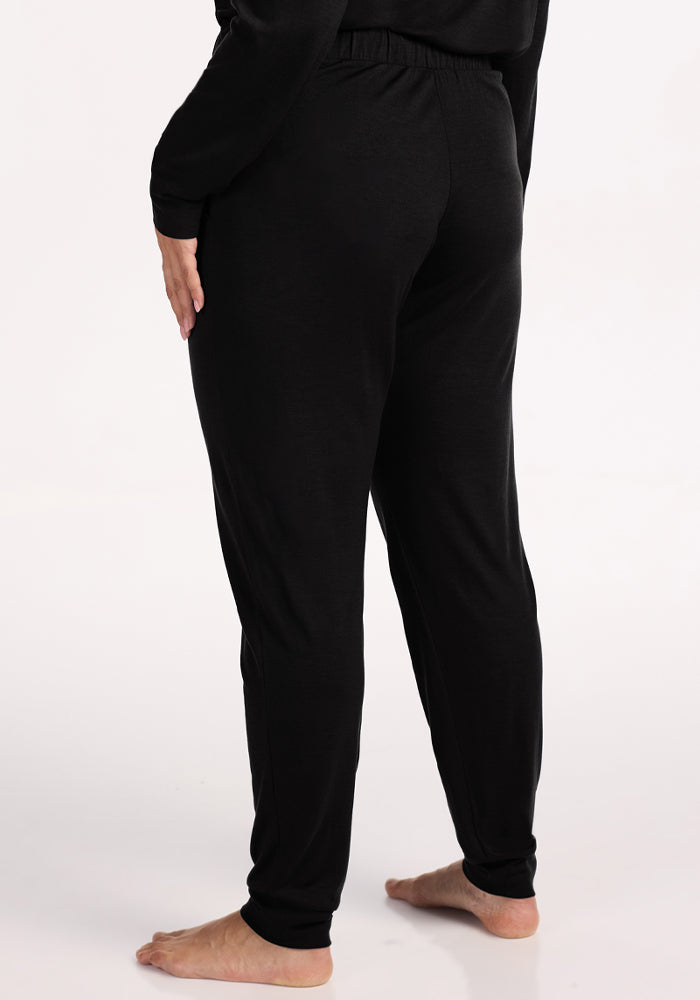 A person stands barefoot on a light surface, shown from the back, wearing loose-fitting Woolx Mary Kate Lounge Pants in black with a matching long-sleeve top—ideal travel loungewear for comfort and style.