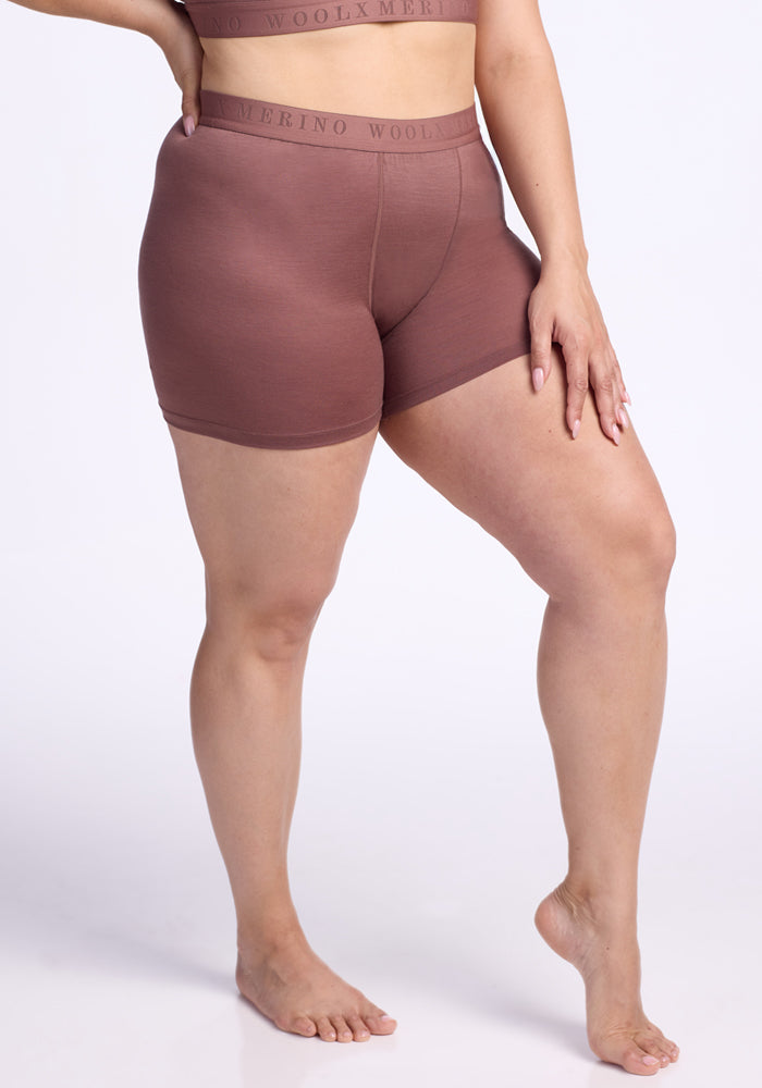A person with medium skin tone wears the Woolx Elliott Boyshort in Nutmeg, paired with a matching top. Barefoot, they pose on one leg with a hand on their thigh against a plain white background. 