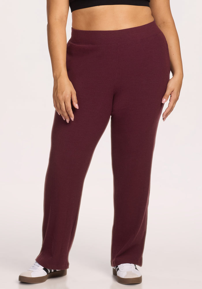 A person in Woolx’s Luca Ribbed Pants in Toasted Garnet, styled with a black crop top and white sneakers, stands against a plain light background. 