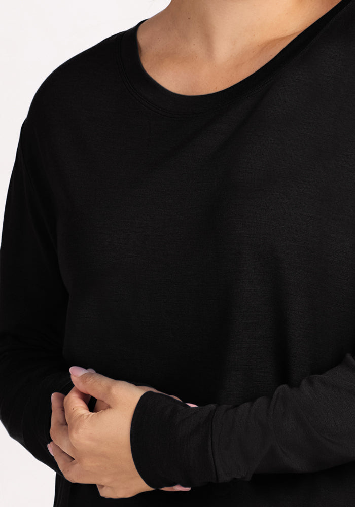 A person wears the Woolx Ashlie Lounge Top - Black, a long-sleeved, wrinkle-resistant Merino wool travel top. They stand with hands clasped at waist level, showing only their upper body and hands against a plain background.