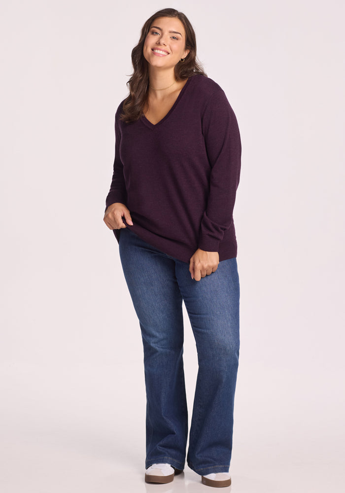 A woman smiles, wearing the Woolx Francesca Sweater in Deep Plum, paired with blue jeans and white sneakers, against a plain light background.