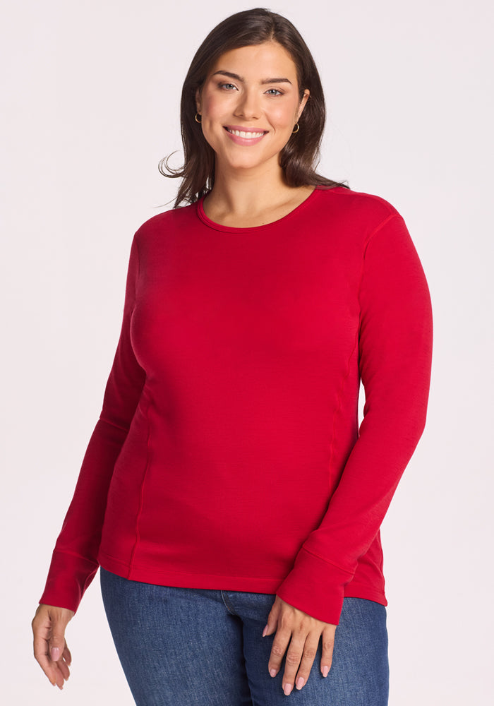 A woman with long brown hair smiles while wearing the Woolx Hannah Long Sleeve Top in Maraschino Cherry and blue jeans. The temperature-regulating, moisture-wicking merino wool keeps her comfortable against a plain light background. 