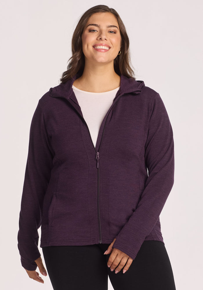 A woman with long brown hair smiles while wearing a Woolx Zoey Zip Up Hoodie in Deep Plum over a white shirt and black pants, standing against a plain light background. 
