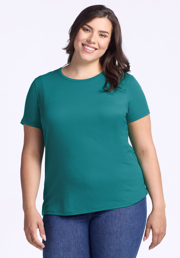 A woman with long brown hair, wearing the Woolx Liza Crew Neck Tee in Surfside and blue jeans, smiles while standing against a plain white background. 