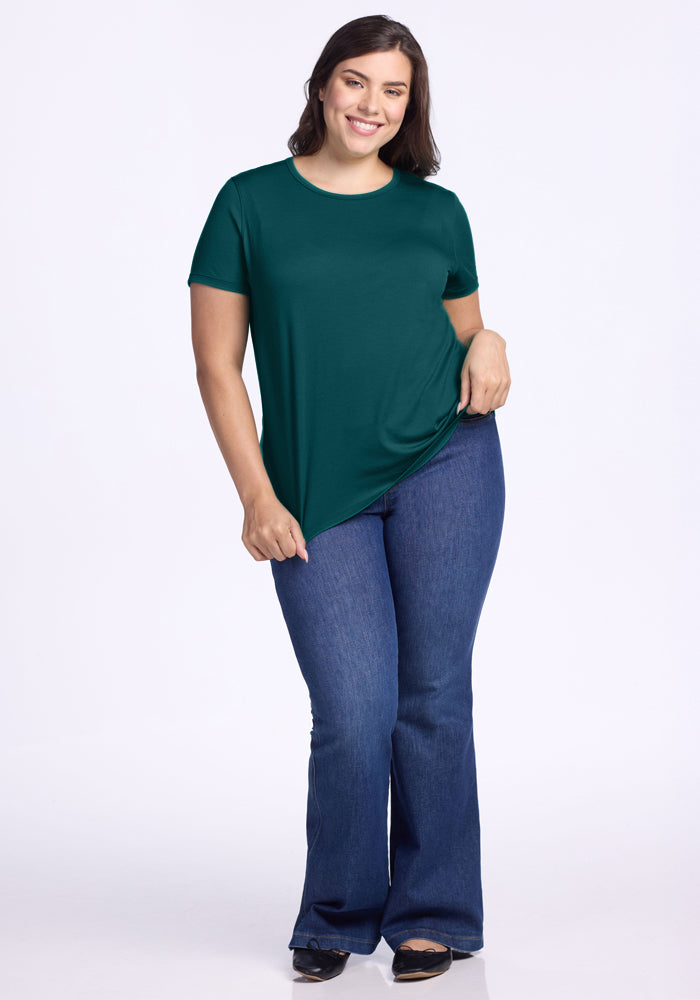 A woman stands smiling against a white background, wearing the Woolx Liza Crew Neck Tee in Riverbank Green and blue flared jeans with black shoes.