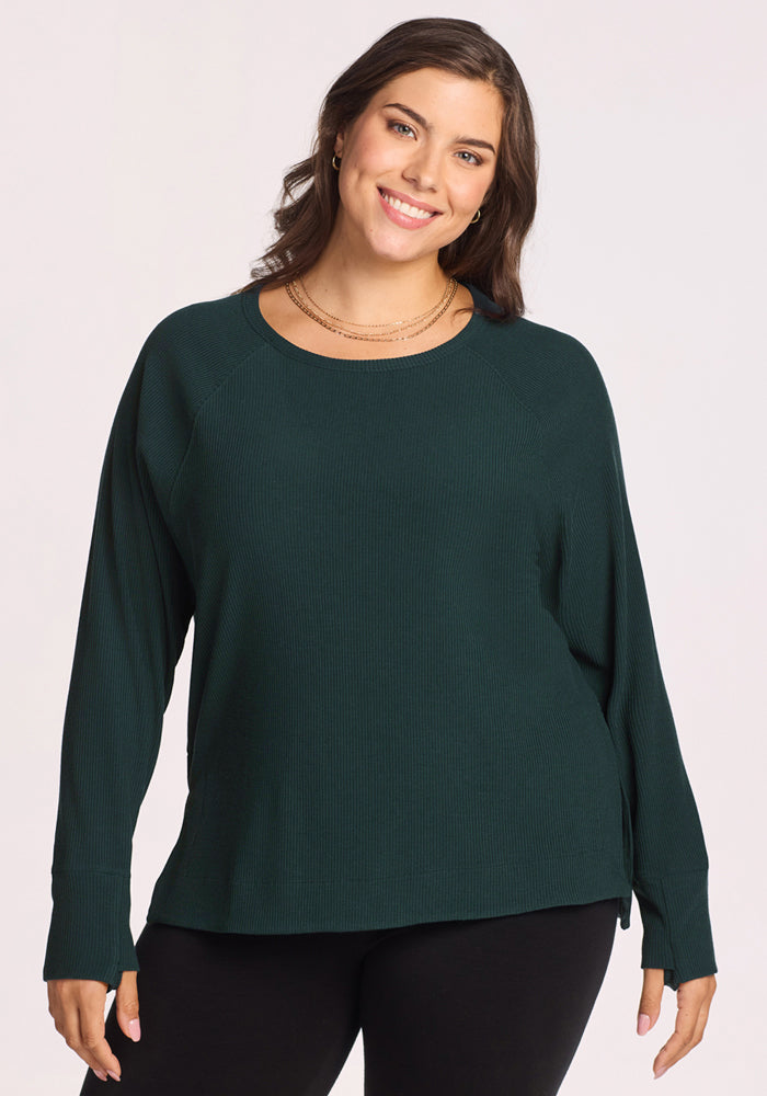 A woman with long brown hair wearing the Woolx Ainsley Sweater in Deep Timber and black pants smiles at the camera against a light background. 