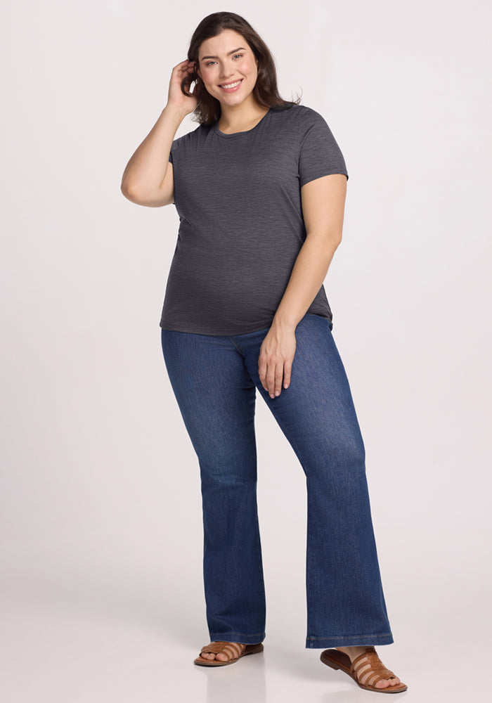 A woman stands smiling and touching her hair, wearing the Woolx Liza Crew Neck Tee in Cinder Melange, blue flare jeans, and brown sandals against a plain light background.