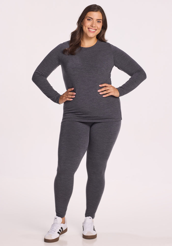 A woman with long dark hair smiles, hands on hips, wearing the Woolx Sadie Baselayer Top in Cinder Melange and matching leggings with white sneakers featuring black stripes, combining comfort and style against a plain white background.