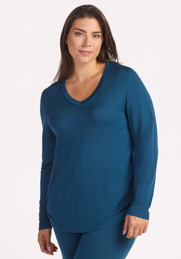 A woman with long brown hair models the Woolx Lily V Neck Lounge Top in Majolica Blue, standing against a white background with a slight smile—ideal comfort for night sweats or as cozy travel sleepwear. 