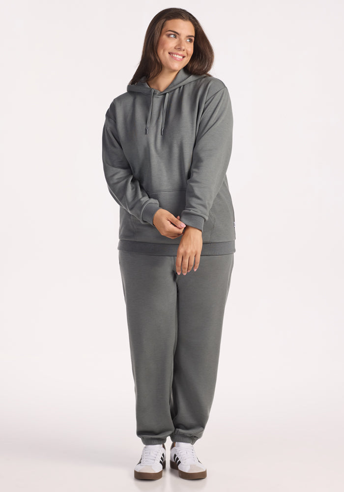 A person stands smiling in a Woolx Avery Hoodie - Sedona Sage and matching sweatpants with white sneakers, against a plain light background.