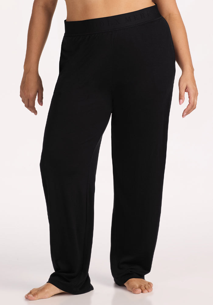 A person models the Woolx Maya Lounge Pants in black, made from Merino wool, standing barefoot against a plain white background with only the lower torso and legs shown. 