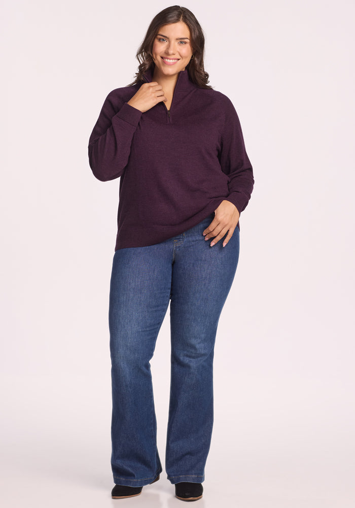 A woman with wavy brown hair smiles while standing in front of a plain light background. She wears the Woolx Collins Half Zip Sweater in Deep Plum, paired with blue jeans and black shoes.