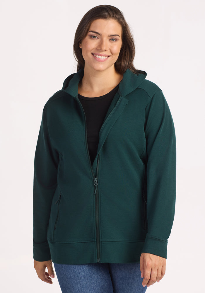 A woman with long brown hair smiles while wearing the Woolx Darby Zip Up Hoodie in Deep Timber over a black shirt and blue jeans, standing against a plain white background. 
