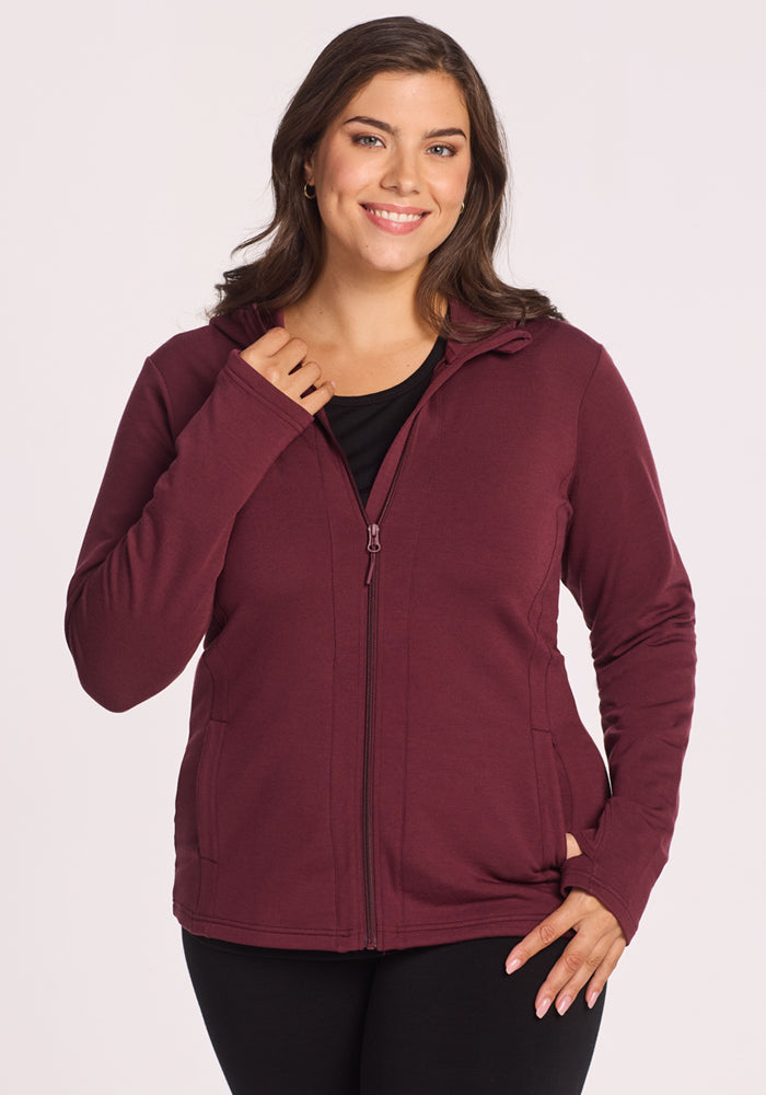 A woman with long brown hair smiles while wearing a Woolx Zoey Zip Up Hoodie in Toasted Garnet over a black top. She stands against a plain background, holding the hoodie slightly open with one hand. 