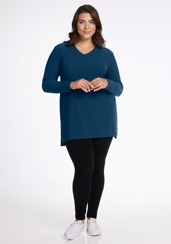 A woman with brown hair smiles, wearing the Woolx Evie Ribbed Tunic in Majolica Blue, paired with black leggings and white sneakers, standing against a plain white background.