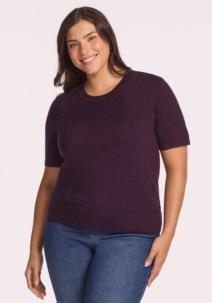 A woman with long brown hair smiles, wearing the Woolx Anna Crew Sweater in Deep Plum and blue jeans, standing against a plain light background. 