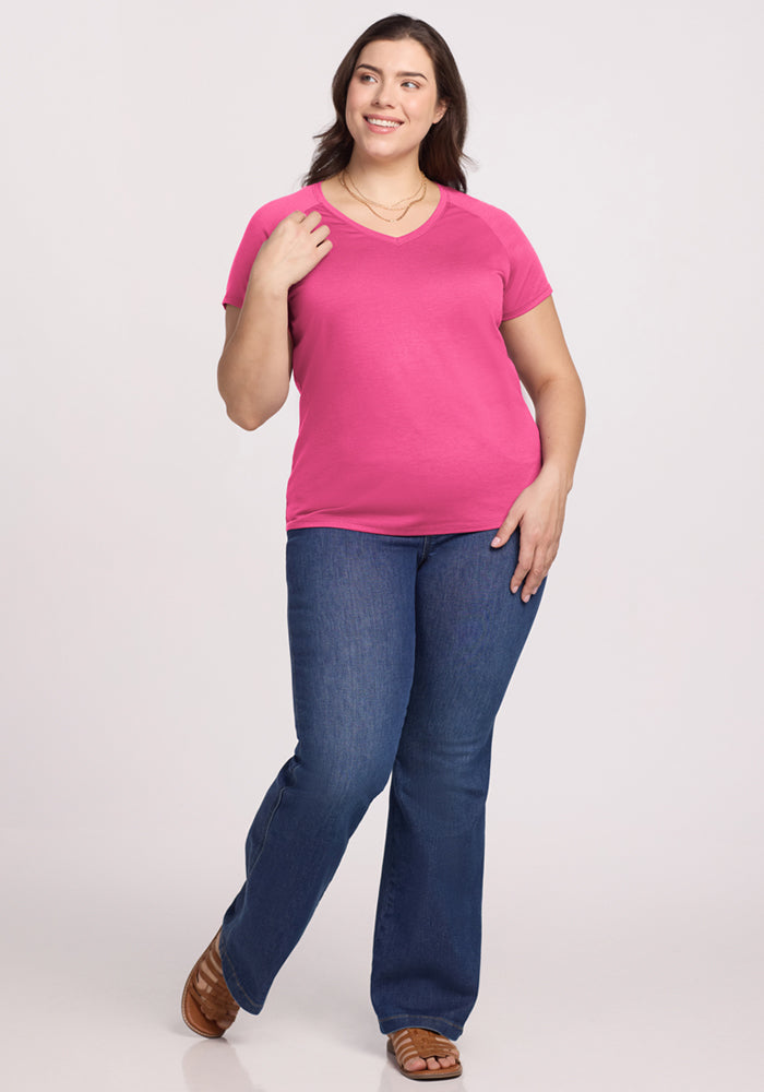 A person with long dark hair is standing and smiling, wearing the Woolx Mia V Neck in Raspberry Fizz, blue jeans, and brown sandals against a plain light background.