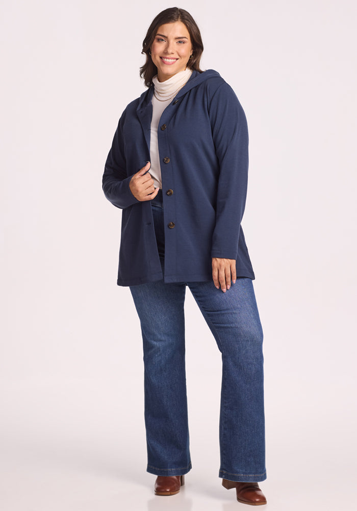 A woman with medium brown hair smiles wearing the Woolx Edna Hooded Cardigan in Deep Navy, paired with a white turtleneck, blue jeans, and brown boots—her ideal cold-weather look against a light background.