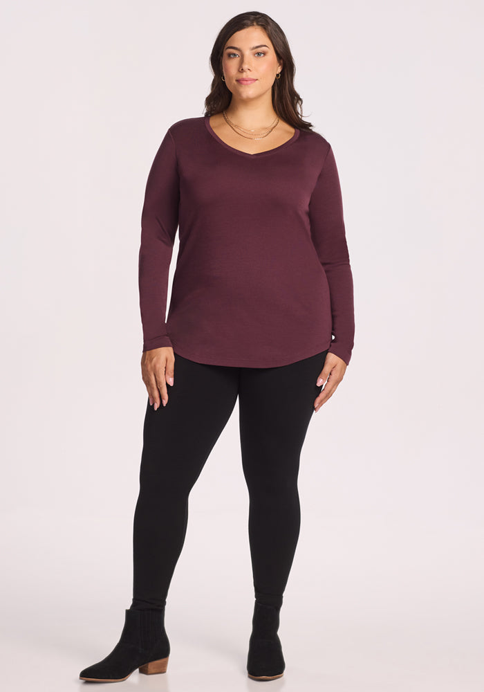A woman with long brown hair wears the Woolx Eva Tunic in Chocolate Fig, black leggings, and black ankle boots while standing against a plain light background.