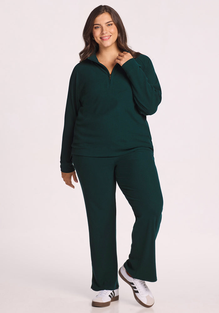 A woman with long dark hair smiles while wearing the Woolx Quinn Ribbed 1/4 Zip in Deep Timber and matching pants with white sneakers, standing against a plain white background.