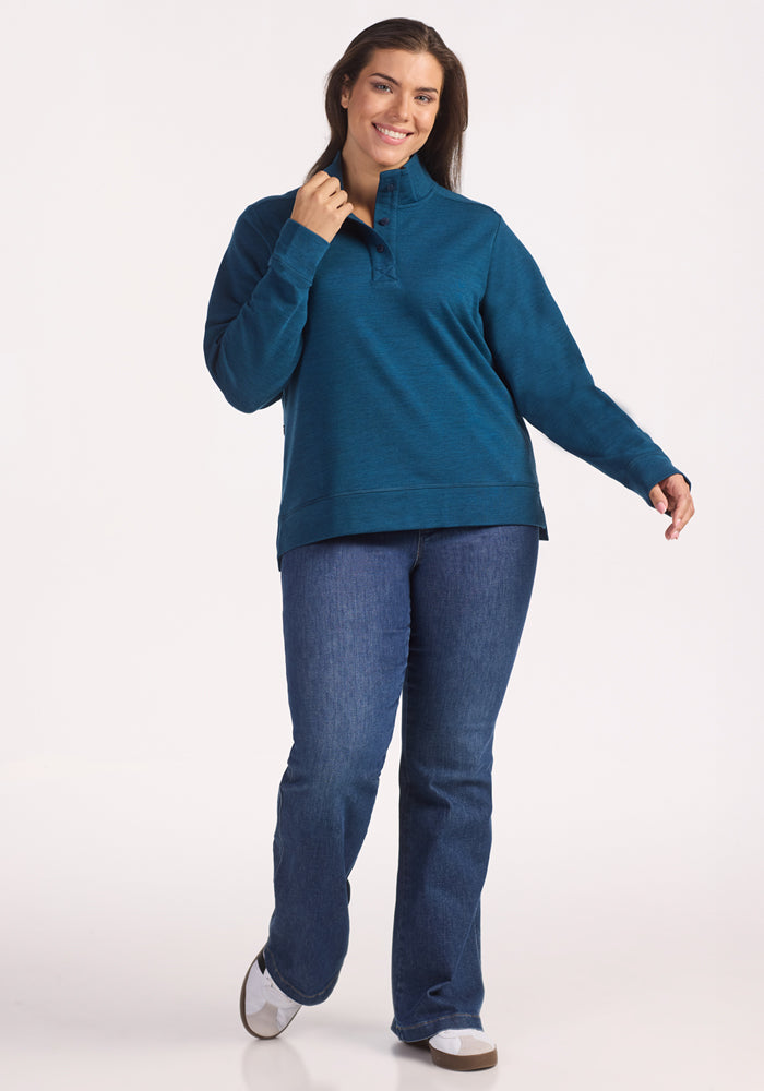A woman with long brown hair smiles against a white background, wearing the Woolx Annie 3 Button Pullover in Majolica Blue, blue jeans, and white shoes. She holds the pullover collar with one hand and steps forward looking confident and relaxed.
