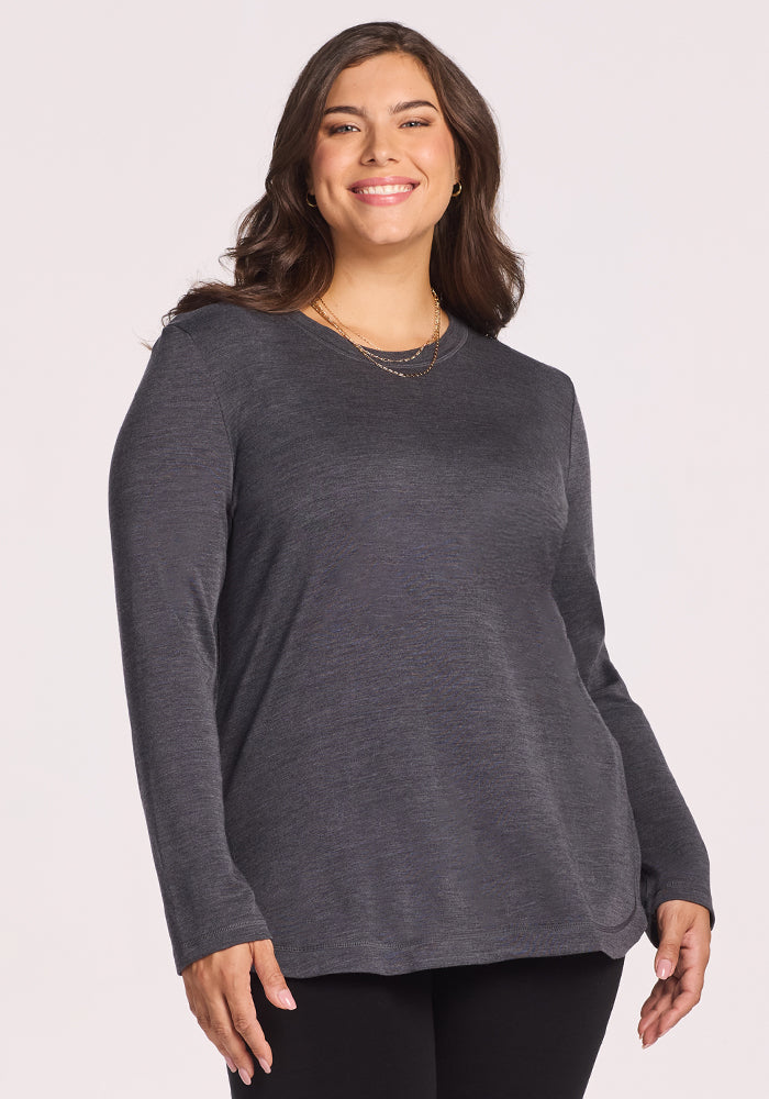 A woman with long brown hair smiles and stands against a light background, wearing Woolx’s Blair Tunic in Cinder Melange and black pants, enjoying its lightweight warmth and odor-resistant comfort. 