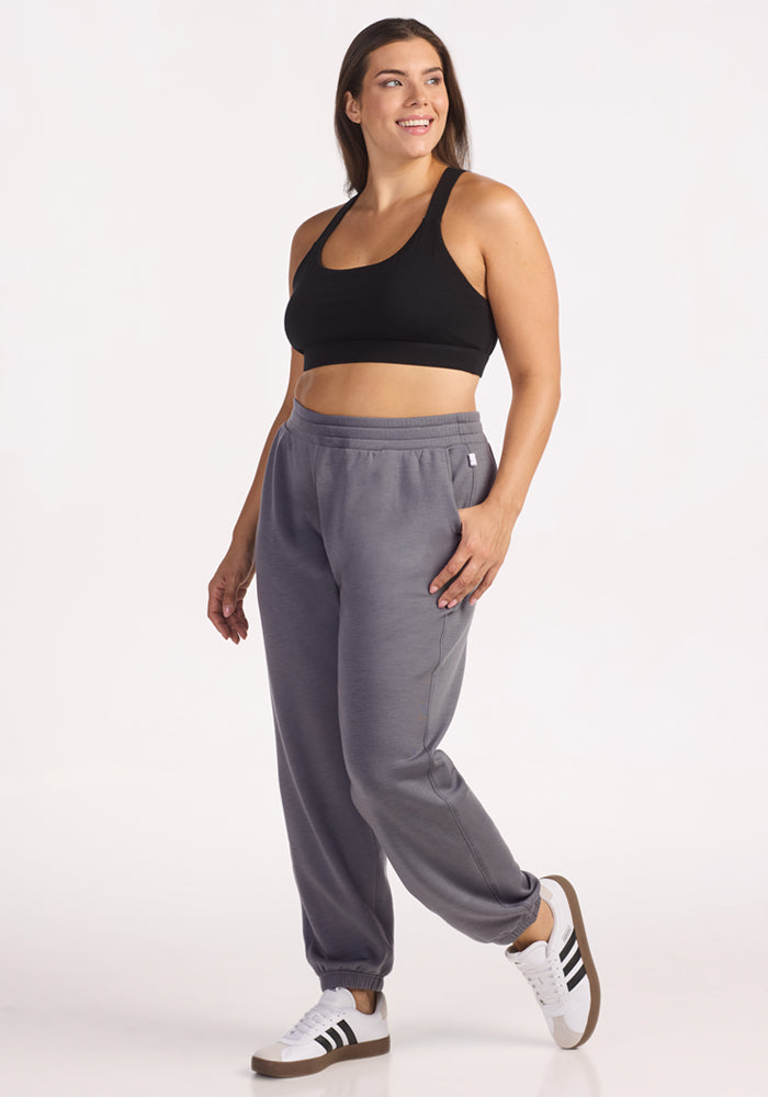 A woman wearing Woolx Parker Sweatpants in Arctic Dusk, a black sports bra, and white sneakers with black stripes stands smiling with hands in pockets, looking to the side against a plain white background.