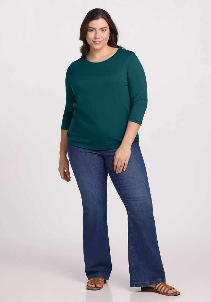 A woman with long dark hair wears the Woolx Jenny 3/4 Sleeve Crew Neck in Riverbank Green, blue flared jeans, and brown sandals. She stands facing forward and smiles softly against a plain light background.
