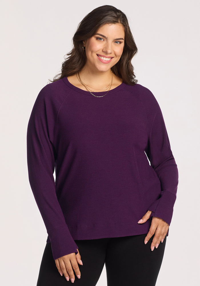 A woman with long brown hair smiles while wearing the Woolx Ainsley Sweater in Majestic Purple and black pants. She stands against a light background, one hand on her hip, highlighting this travel-friendly, temperature-regulating style. 