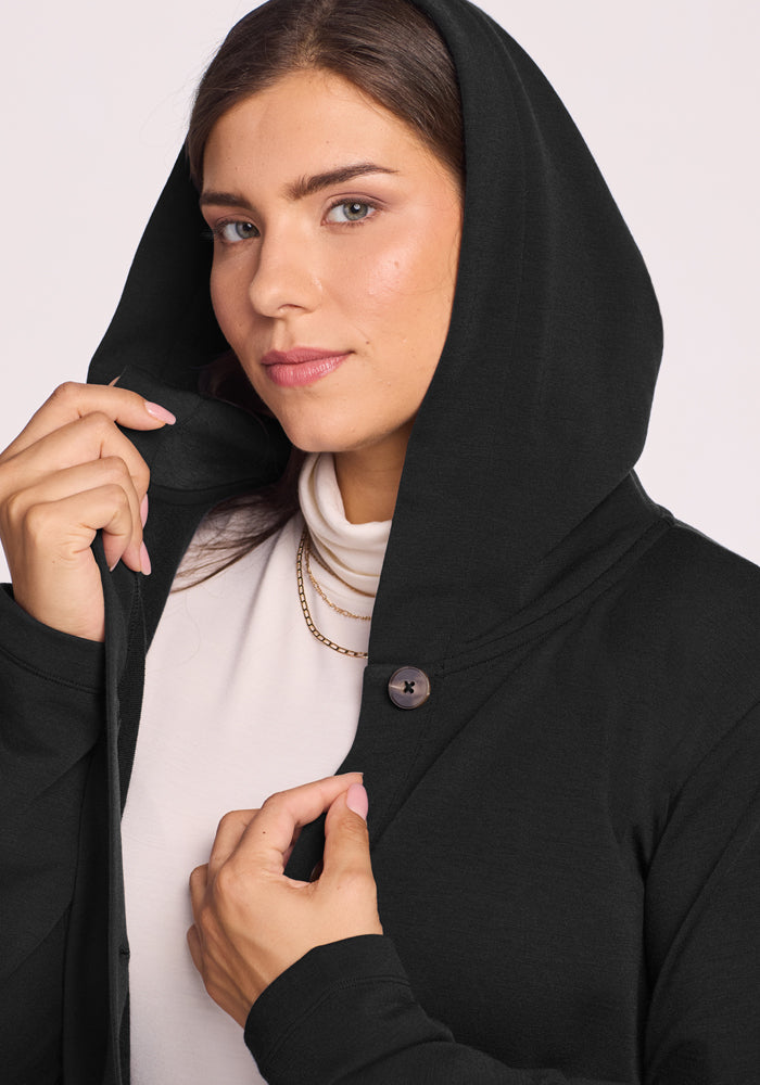 A brunette woman wears the Woolx Edna Hooded Cardigan in black over a white top, softly holding the hood by her face. A button and gold necklace add elegance to her cozy cold-weather look.