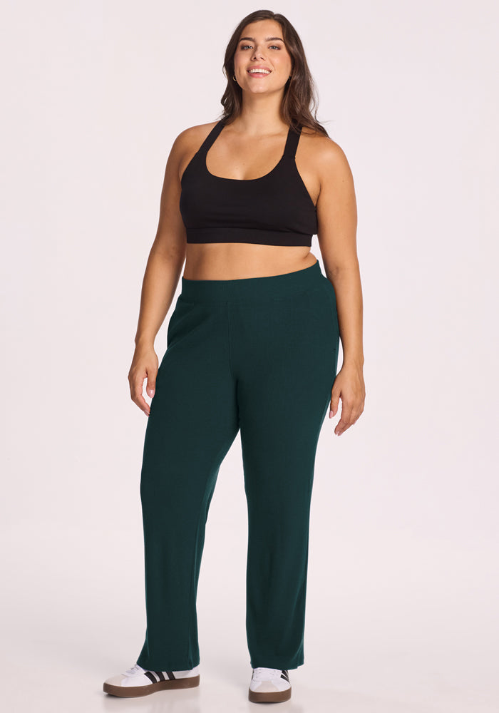 A woman with long brown hair smiles while wearing Woolx's Luca Ribbed Pants in Deep Timber, paired with a black sports bra and white sneakers with black stripes, standing against a plain light background.