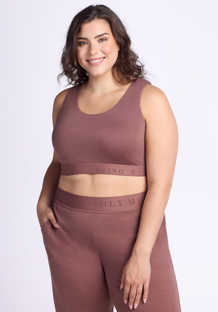 A woman with wavy brown hair smiles in a mauve Woolx Aubrey Lounge Bra - Nutmeg and matching high-waisted pants, one hand in her pocket, against a plain light background.