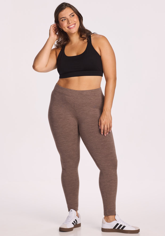 A woman in Woolx Stella Leggings in Simply Taupe, a black sports bra, and white Adidas sneakers stands smiling with one hand touching her hair against a plain white background.