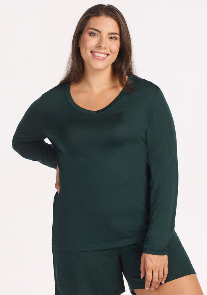 A woman with long brown hair, wearing the Woolx Lily V Neck Lounge Top in Deep Timber, stands smiling with one hand on her hip against a plain white background. 