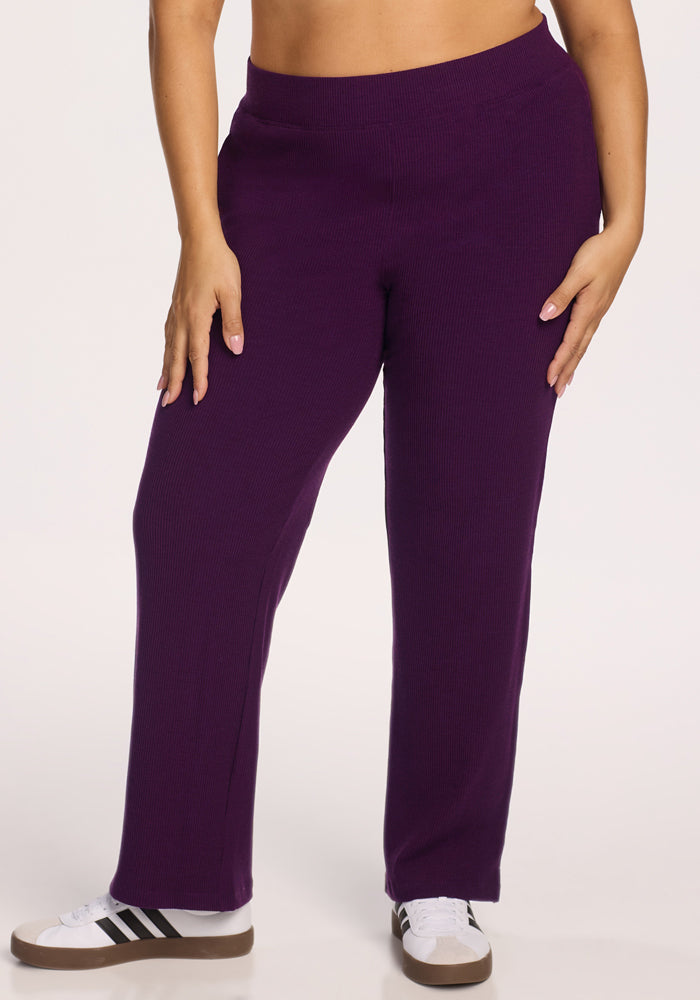 A person wearing Woolx Luca Ribbed Pants in Majestic Purple and white sneakers with black stripes stands against a plain light background. 