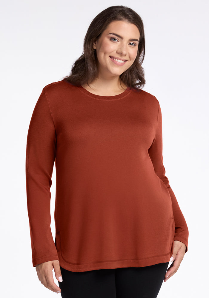 A woman with long brown hair smiles while wearing the Woolx Blair Tunic in Maple Leaf and black pants, standing against a plain white background. 