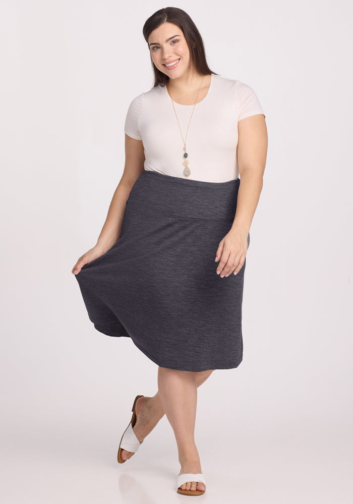 A woman with long brown hair wears a white top, white sandals, and the Woolx Marilyn Flowy Midi Skirt in Cinder Melange. She smiles, posing with one hand holding her skirt against a plain light background.